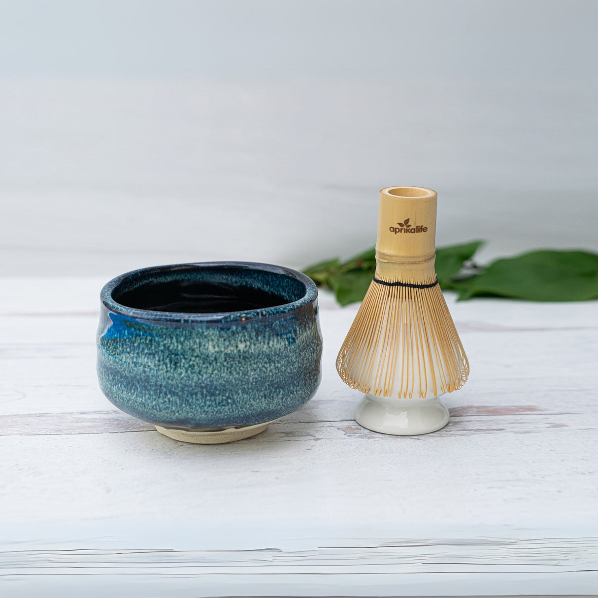 The Maruyoshi Navy Sea Ceramic Matcha Bowl and a bamboo whisk sit elegantly on a white surface, framed by green leaves, creating a serene backdrop for this exquisite Matcha Tea Set.