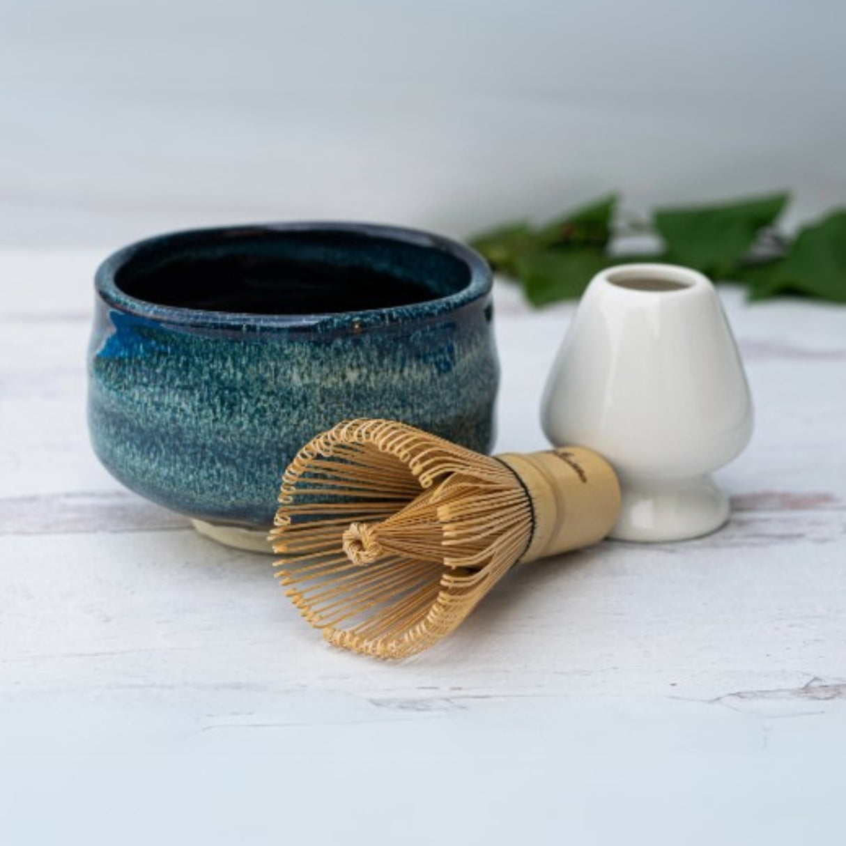 The Maruyoshi Navy Sea Ceramic Matcha Bowl with a Bamboo Matcha Whisk and white holder rests on a light surface, framed by a green leaf backdrop.