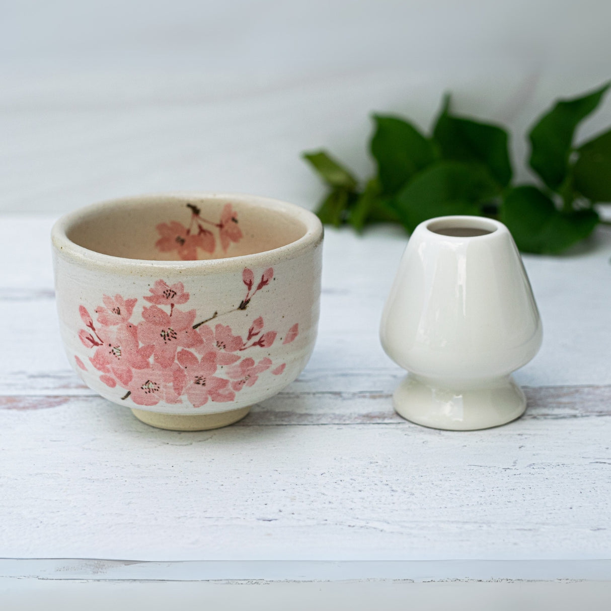 Maruyoshi Sakura Clay Matcha Bowl, a bamboo whisk with holder, and organic Japanese matcha set on a white wooden surface with green leaves in the background.