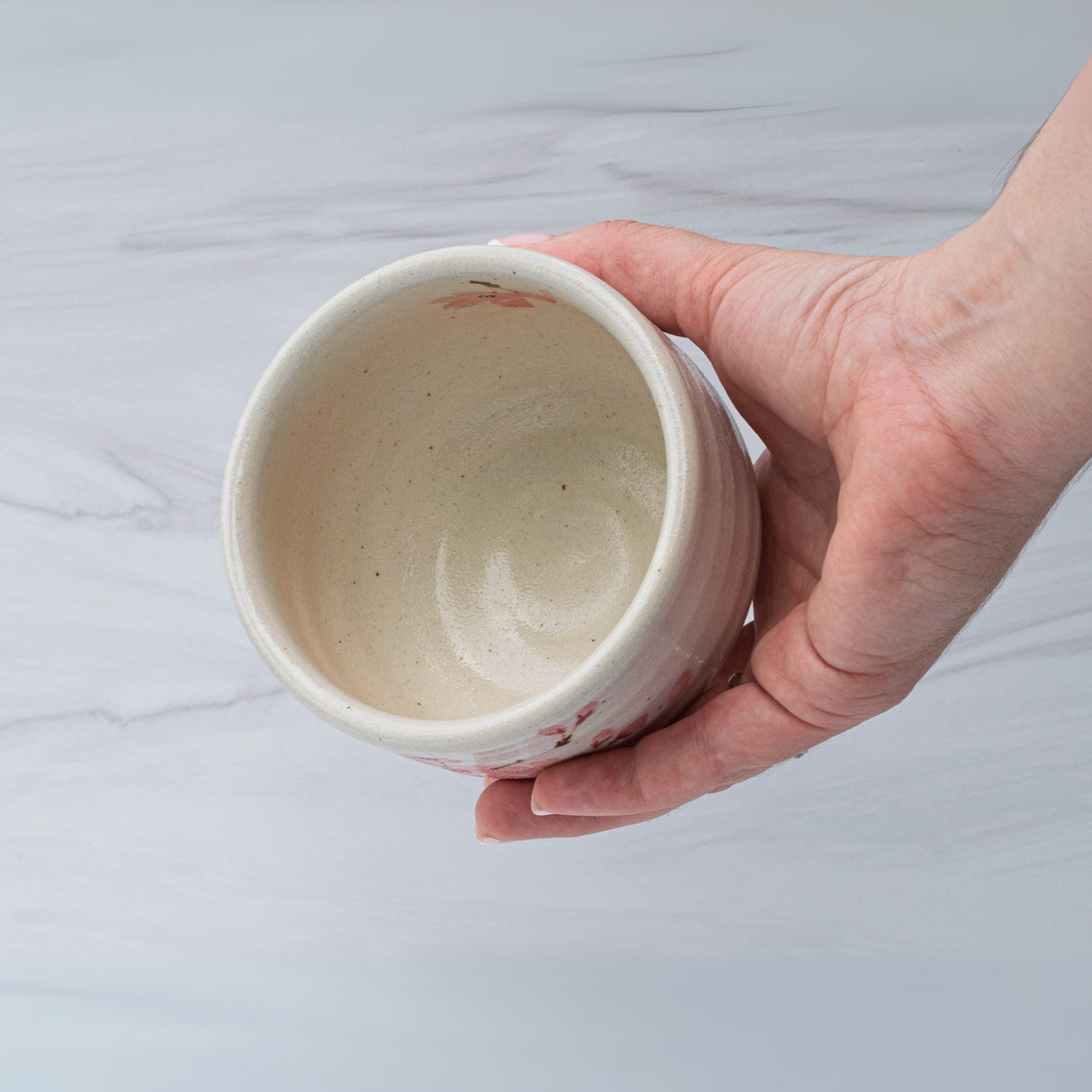 A hand cradles a Maruyoshi Sakura Clay Matcha Bowl, part of the set with a Bamboo Whisk and Organic Japanese Matcha, against a marble-like background.