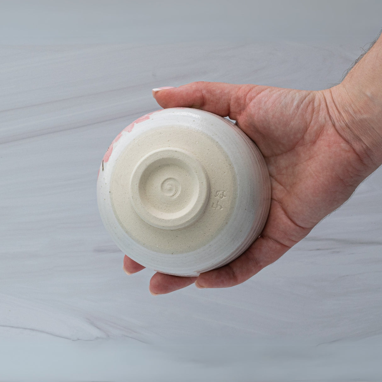 A hand holds an upside-down Maruyoshi Sakura Clay Matcha Bowl, showcasing a spiral design against a light background. This bowl, crafted with precision and elegance, reflects the timeless art of Japanese pottery.