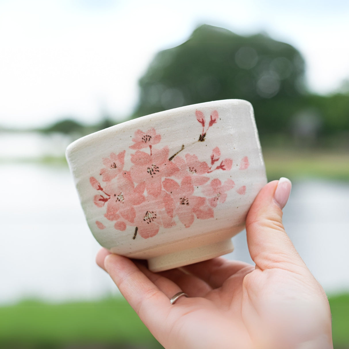A hand elegantly holds a Maruyoshi Sakura Clay Matcha Bowl, adorned with pink cherry blossoms, set against a blurred natural background.