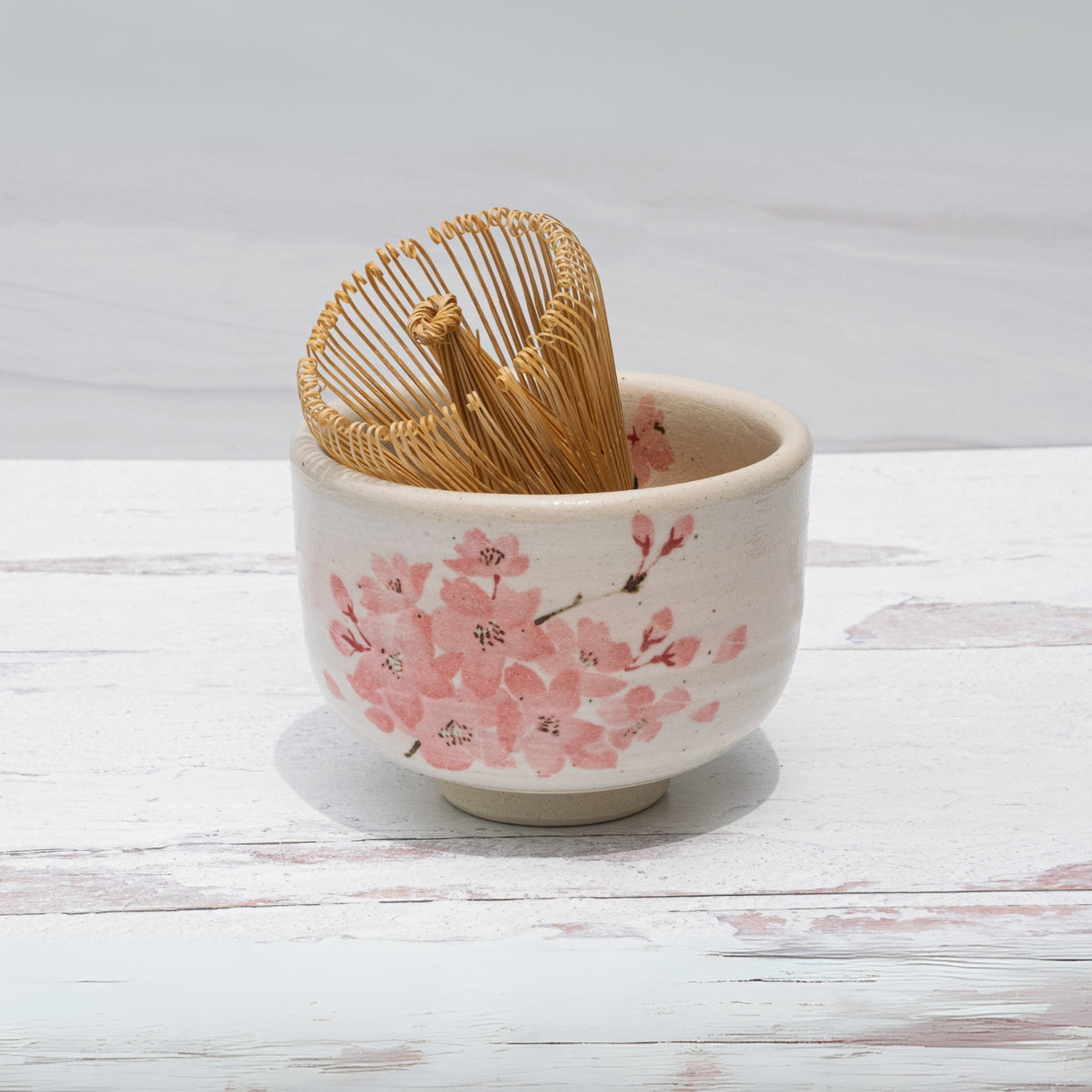 Maruyoshi Sakura Clay Matcha Bowl with cherry blossoms and a Bamboo Matcha Whisk are elegantly arranged on a white wooden surface.