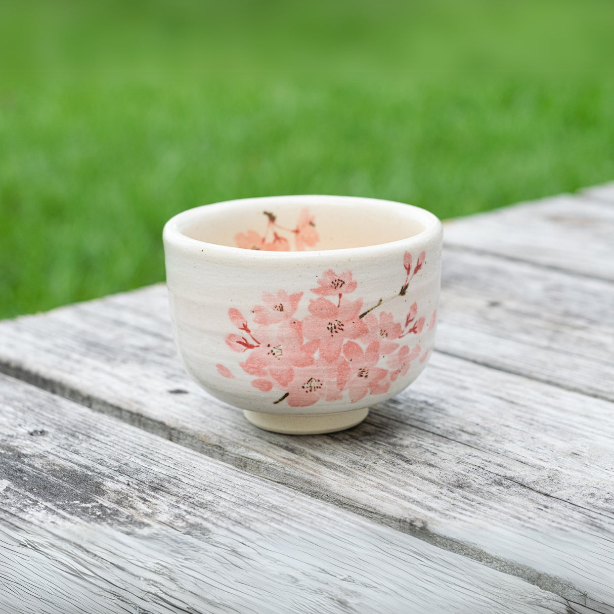 On a weathered wooden surface, the Maruyoshi Sakura Clay Matcha Bowl features pink flowers, set against a backdrop of lush green grass.