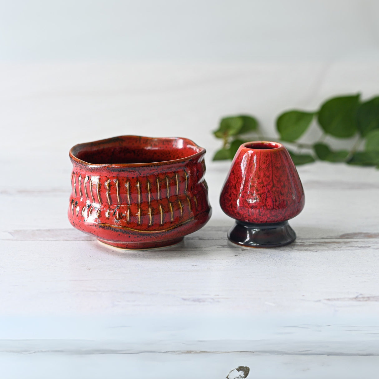 A Meisen Red Matcha Bowl and small vase sit on wood, reflecting Minoyaki elegance. Green leaves enhance the tranquil scene like a traditional Japanese matcha bowl setting, featuring the Bamboo Matcha Whisk, Whisk Holder, and Organic Japanese Matcha Set.