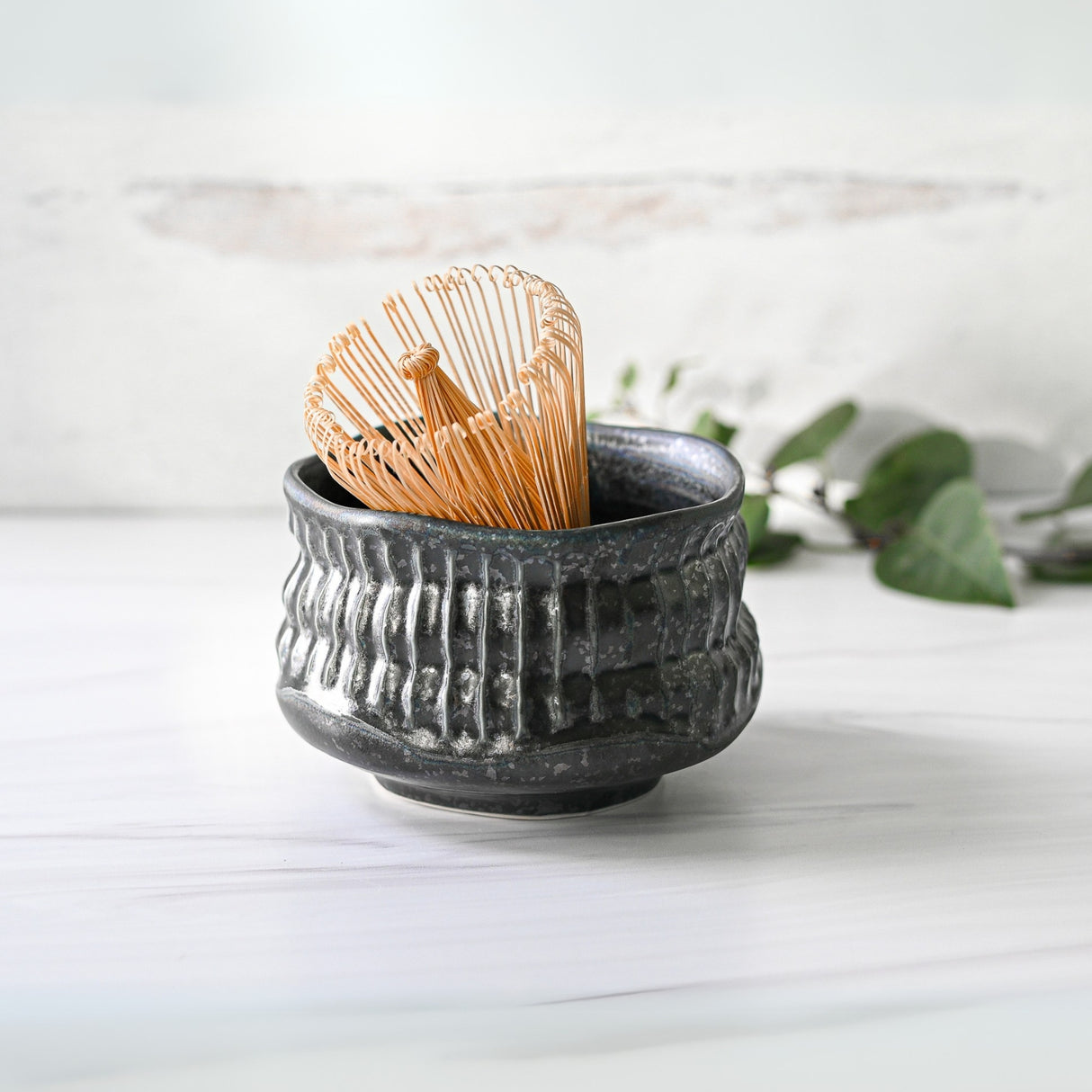 The Black Ceramic Meisen Matcha Bowl, Bamboo Matcha Whisk, Whisk Holder and Organic Japanese Matcha Set rests on a white surface, with softly blurred green leaves in the background.