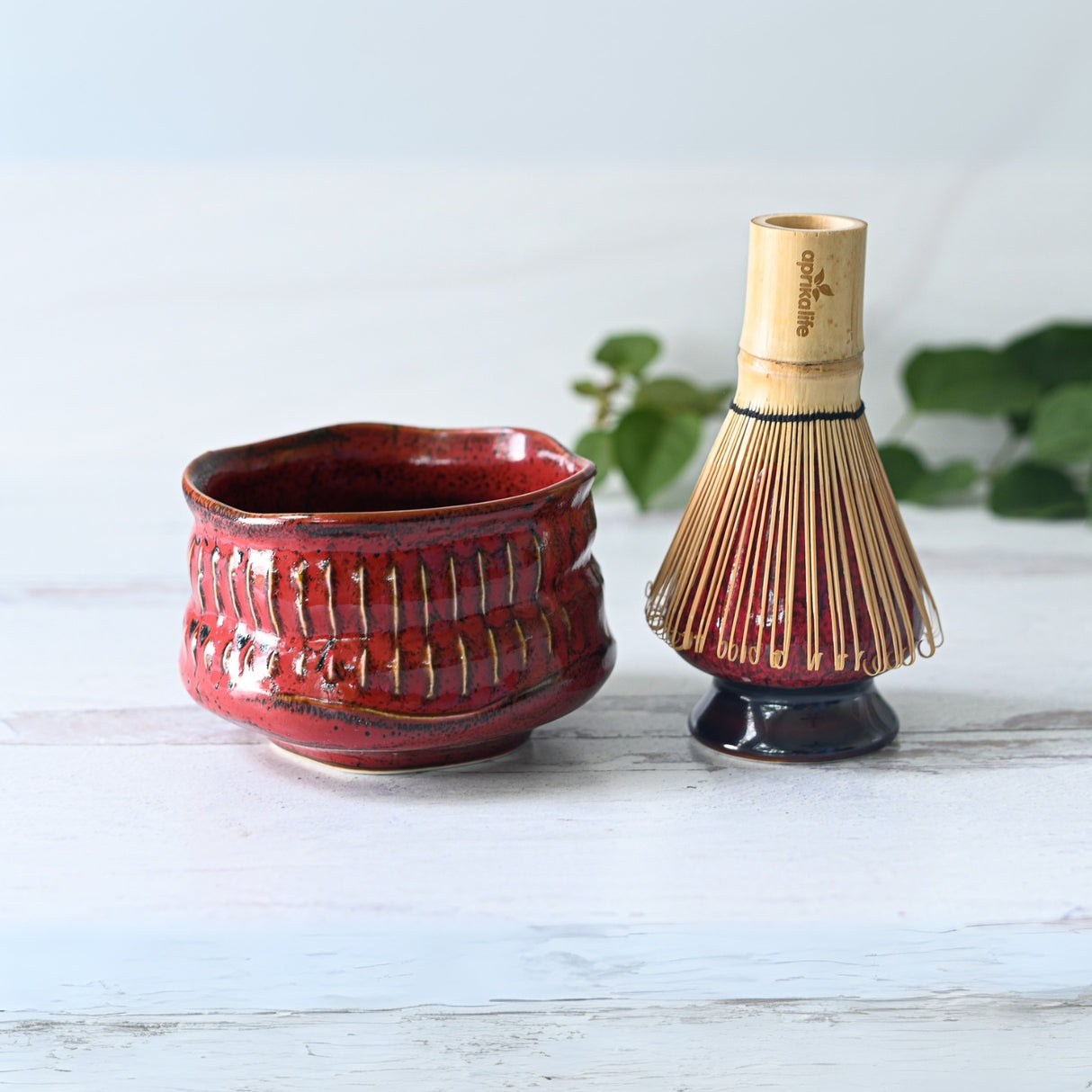 A Meisen Red Matcha Bowl and Bamboo Matcha Whisk grace a light surface, accompanied by green leaves that gently frame the scene.