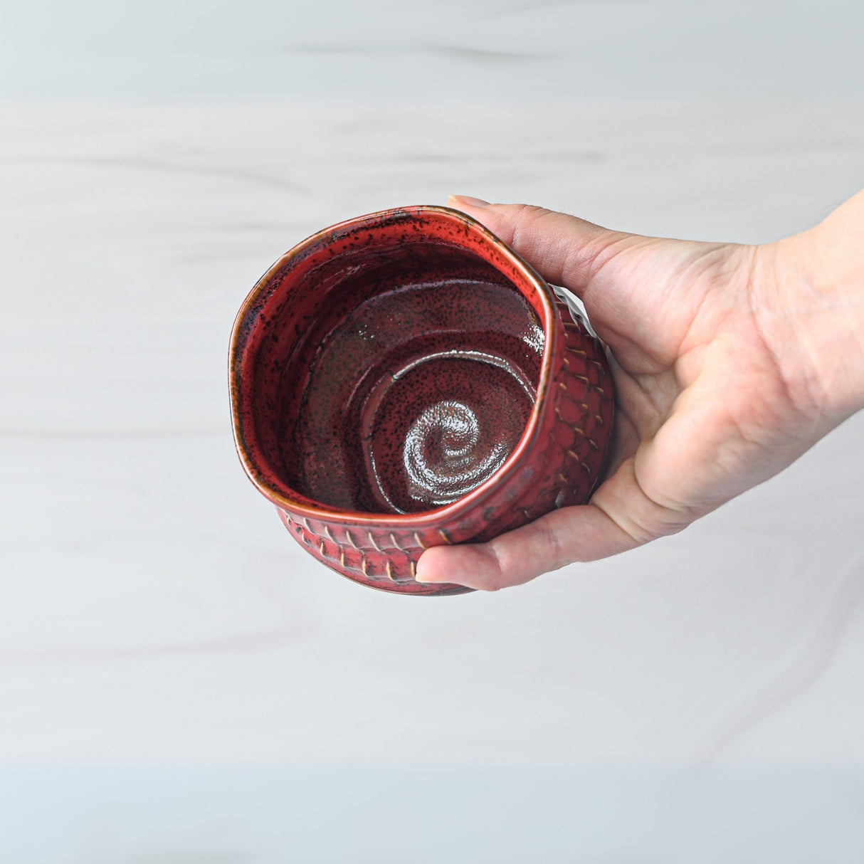 A hand cradles a Meisen Red Matcha Bowl, its red, textured ceramic exterior revealing a captivating spiral pattern inside, reminiscent of Minoyaki craftsmanship.