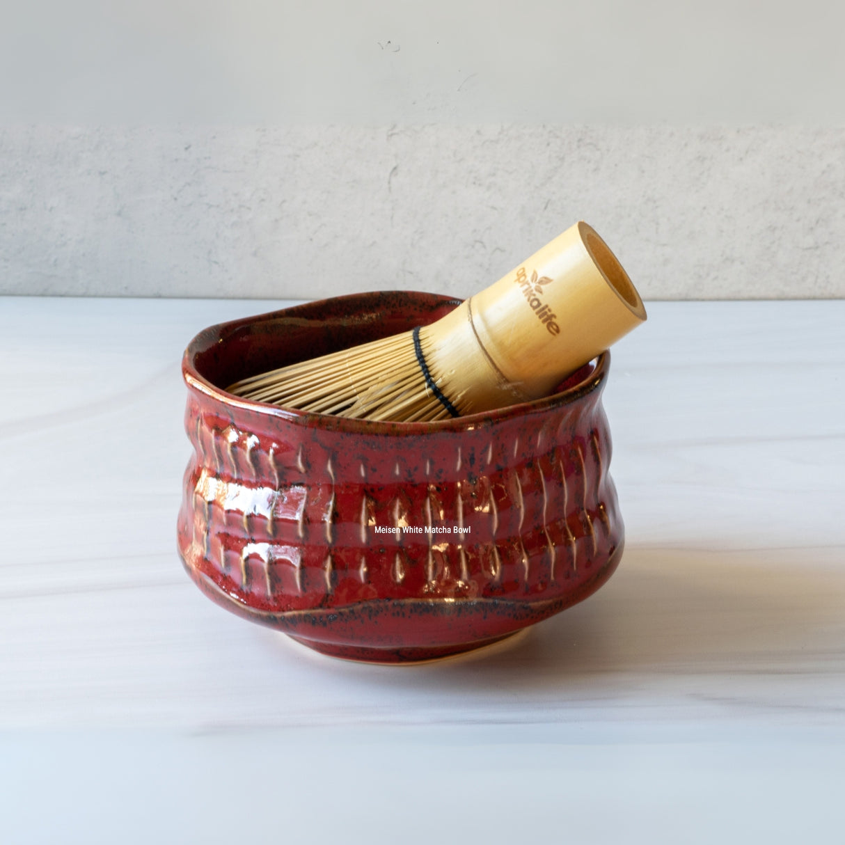 The Meisen Red Matcha Bowl holds a bamboo whisk, beautifully positioned on a light marble surface.