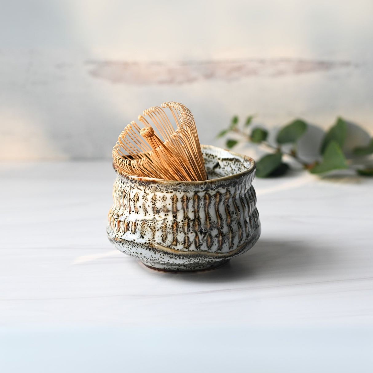 The White Ceramic Meisen Matcha Bowl and Bamboo Matcha Whisk with holder are elegantly set on a white surface, complemented by delicate eucalyptus leaves in the background.