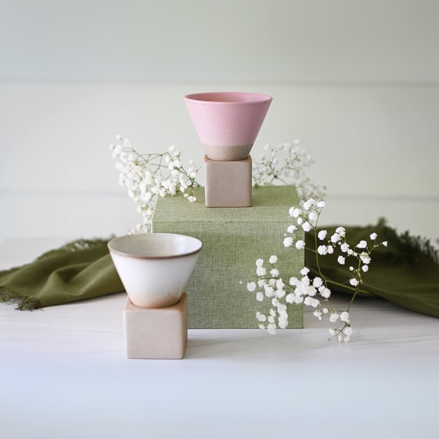 Two vintage-style pink ceramic mugs on square stands with white flowers and green cloth in a minimalist setting.