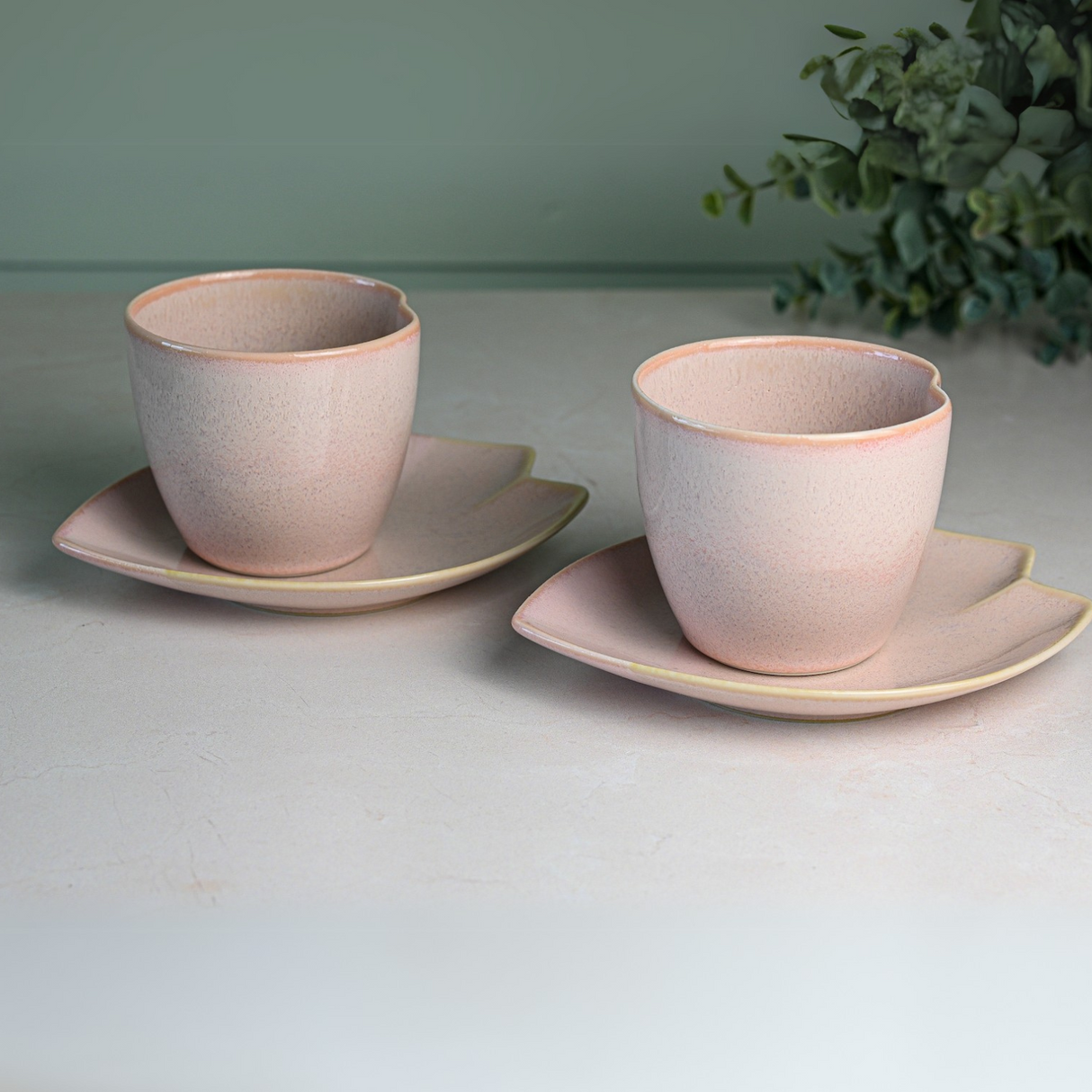 Two Pink LOVE Ceramic Tea Cups with saucers are placed on a light surface, surrounded by leafy greenery in the background.