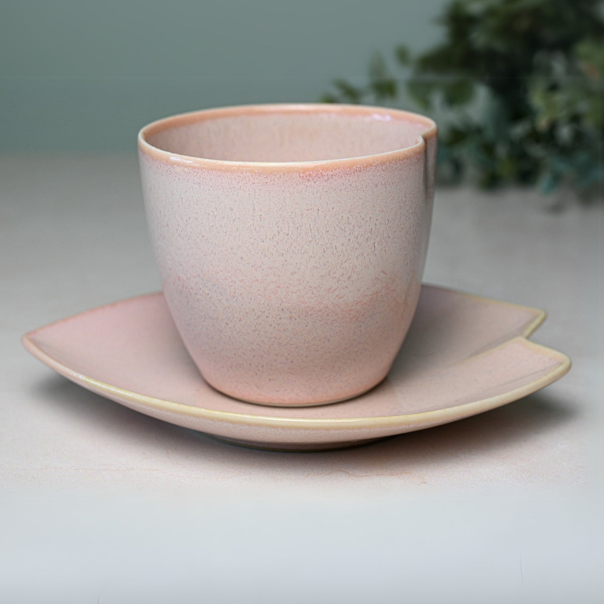 A Pink LOVE Ceramic Tea Cup with Saucer sits against a blurred leafy backdrop, showcasing elegant charm.