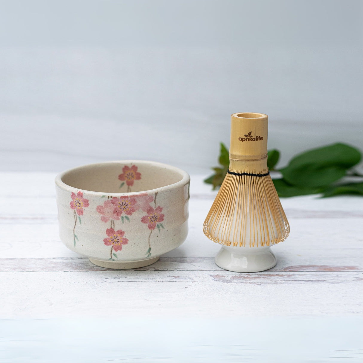 On a white surface lies the Pink Sakura Flowers Ceramic Matcha Bowl with a bamboo matcha whisk, part of an exquisite set including a whisk holder and organic Japanese matcha.