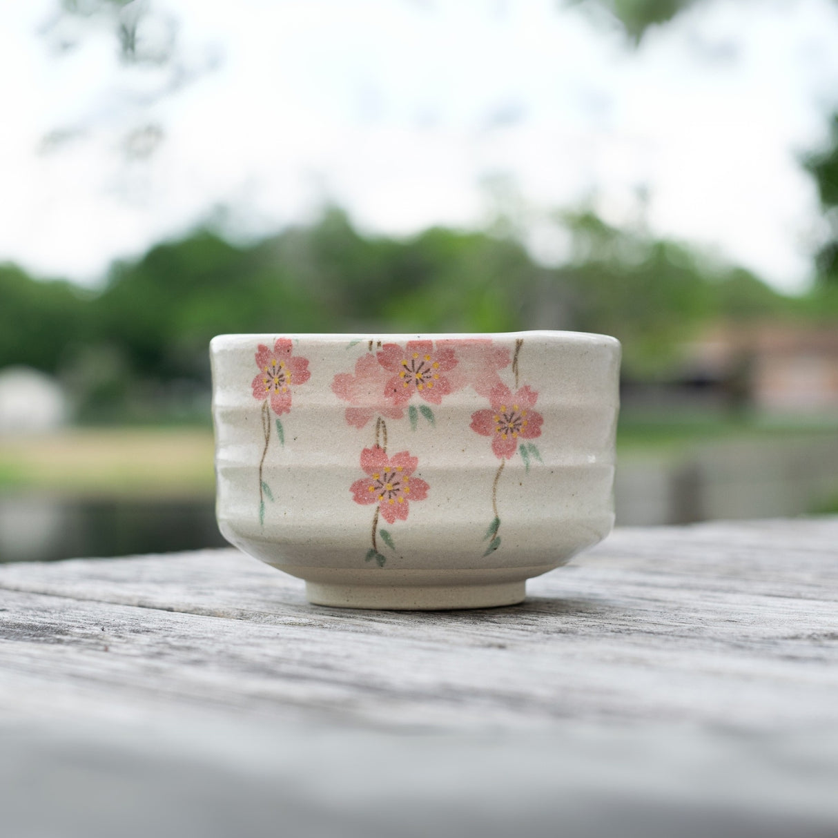 The Pink Sakura Flowers Ceramic Matcha Bowl from the Japanese pottery matcha accessory set is elegantly placed on a wooden table with blurred greenery in the background.