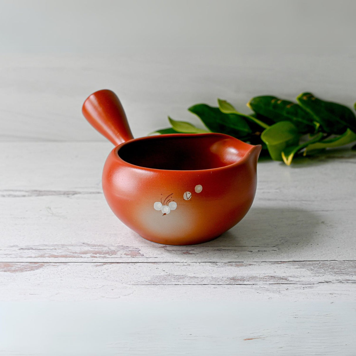 A Red Flowers Ceramic Matcha Bowl with Spout, accompanied by a bamboo matcha whisk and holder, sits on a white wooden surface. Green leaves in the background enhance the tranquil vibe, reminiscent of a Japanese tea ceremony.