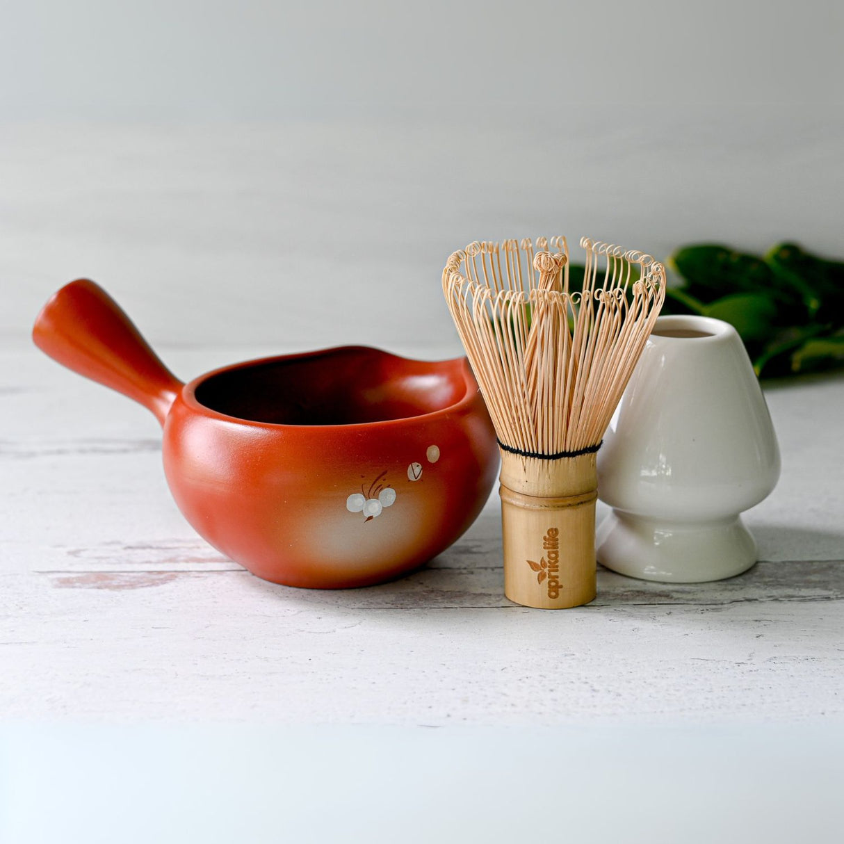 The Red Flowers Ceramic Matcha Bowl with Spout, Bamboo Matcha Whisk, and Whisk Holder on a light wooden surface evoke the essence of a Japanese tea ceremony.