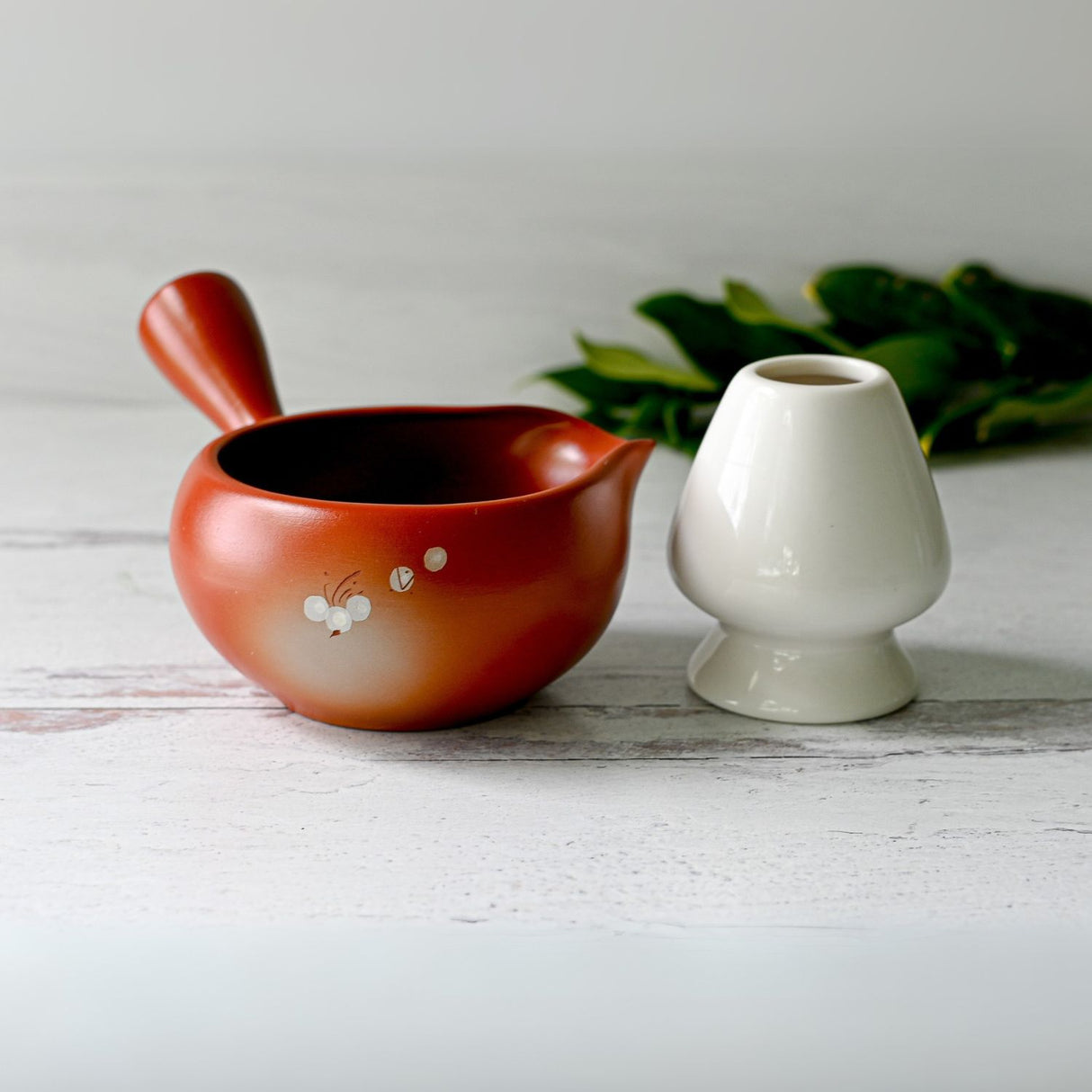 A Red Flowers Ceramic Matcha Bowl with Spout and Bamboo Whisk Set, accompanied by a white vase, sits on a white surface amid green leaves, echoing the calm elegance of a Japanese tea ceremony.
