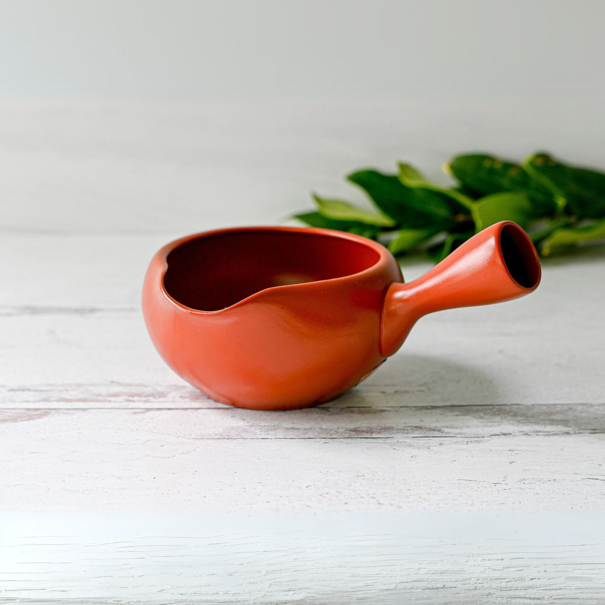 A Red Flowers Ceramic Matcha Bowl with a spout sits on a white wooden surface, surrounded by green leaves, evoking the serene elegance of a Japanese tea ceremony.