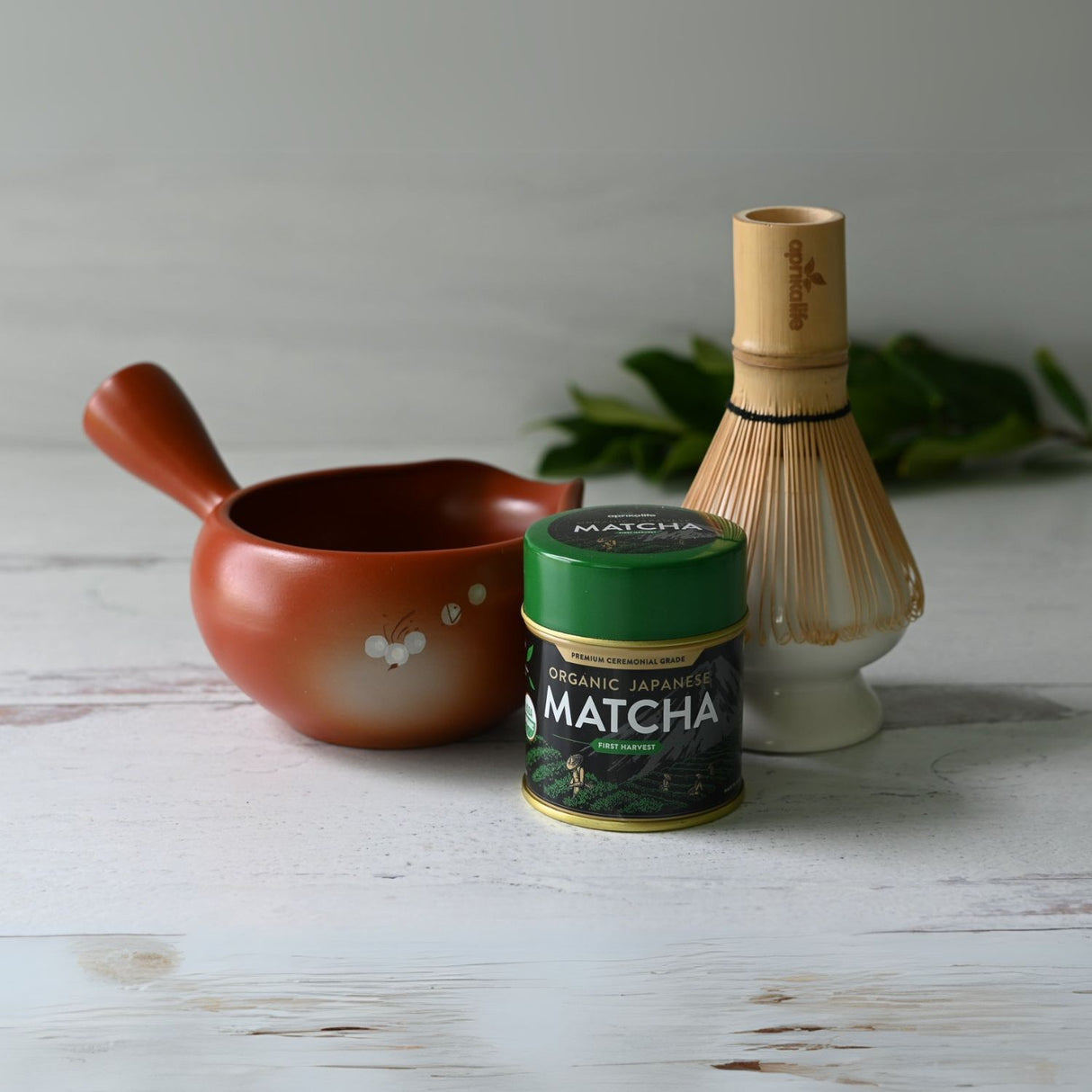 A Red Flowers Ceramic Matcha Bowl with Spout, accompanied by a bamboo matcha whisk, whisk holder, and organic Japanese matcha, sits elegantly on a white wooden surface sprinkled with green leaves, capturing the serene elegance of a Japanese tea ceremony.