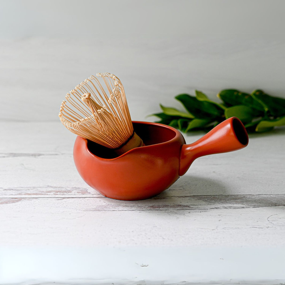 The Red Flowers Ceramic Matcha Bowl with Spout, accompanied by a Bamboo Matcha Whisk and Holder Set, rests on a white wooden surface. Green leaves in the background enhance the serene elegance reminiscent of a Japanese tea ceremony.