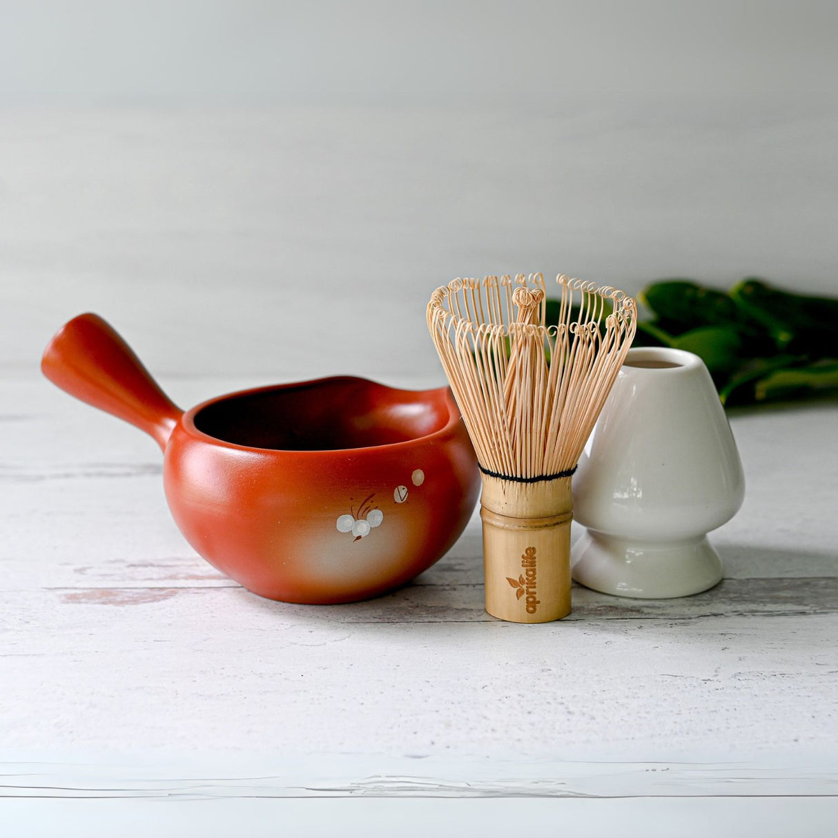The Red Flowers Ceramic Matcha Bowl with Spout, Bamboo Matcha Whisk, and Whisk Holder Set elegantly rests on a light wooden surface, evoking the essence of a Japanese tea ceremony.