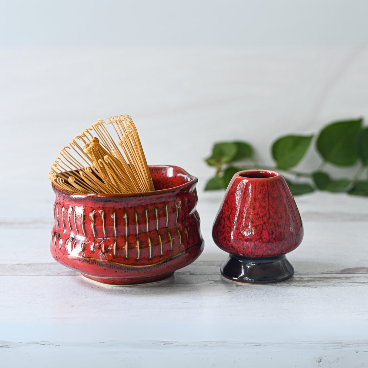 A stunning Red Ceramic Matcha Bowl, paired with a Bamboo Matcha Whisk, Whisk Holder, and Organic Japanese Matcha Set, elegantly rests on a white surface with green leaves in the background.