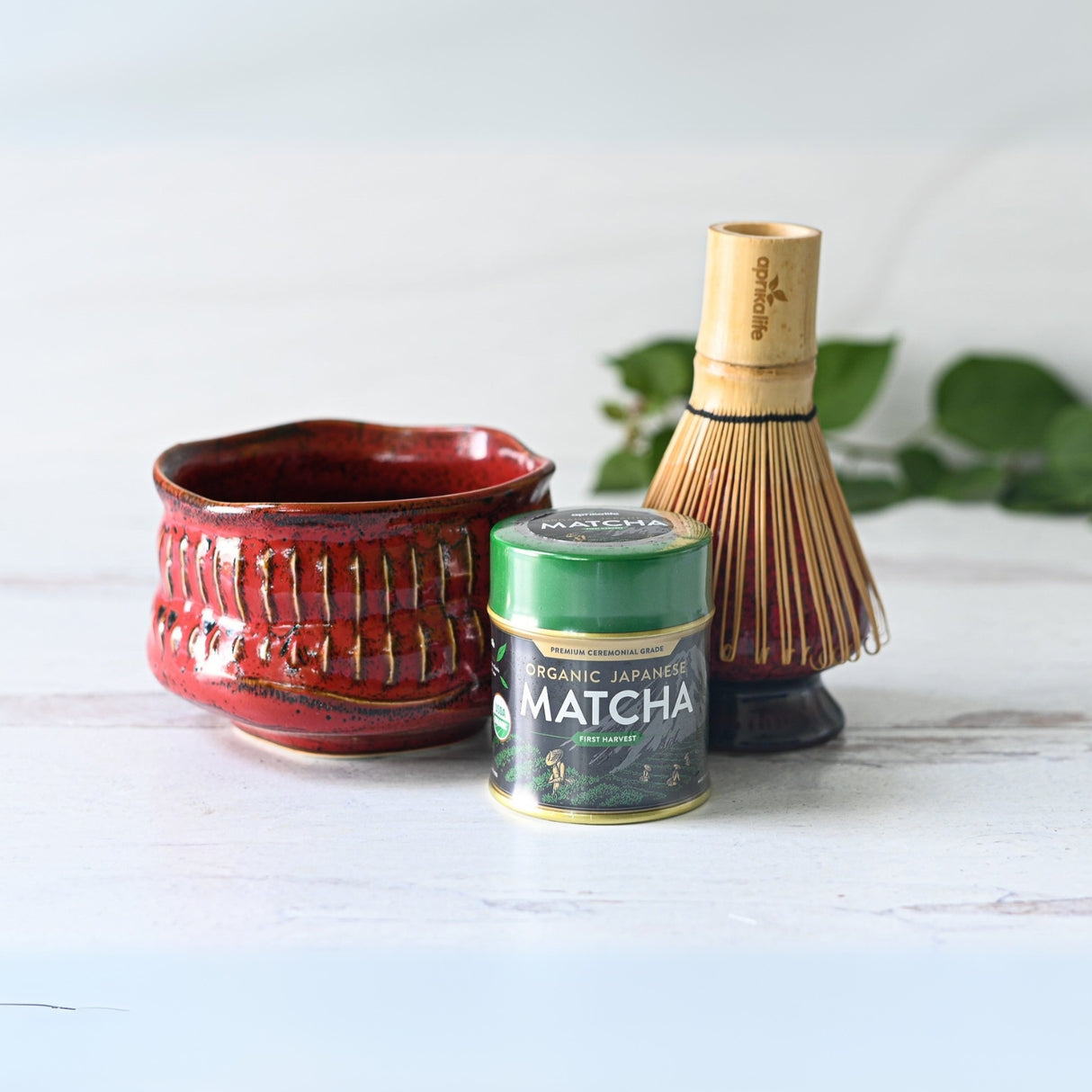 The Red Ceramic Matcha Bowl, Bamboo Matcha Whisk, and Organic Japanese Matcha set are elegantly displayed on a light wooden surface, framed by vibrant green leaves in the background.
