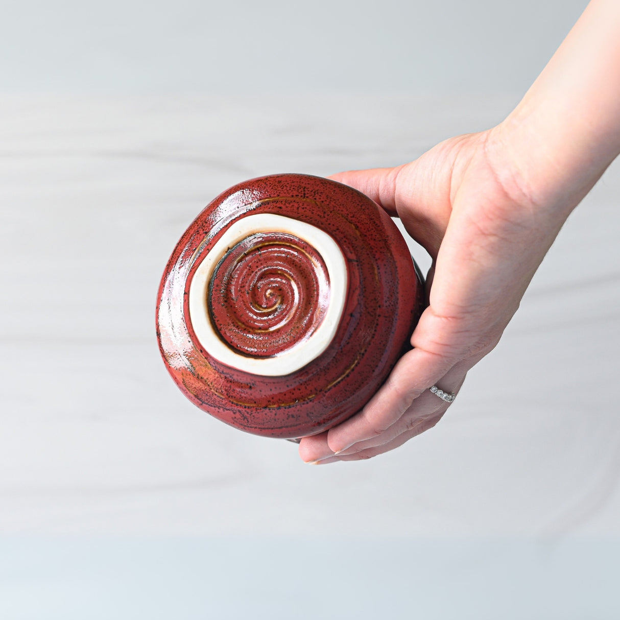 A hand cradles a red Ceramic Matcha Bowl, its bottom adorned with a delicate white spiral design, from the Matcha Set which includes a Bamboo Whisk and Holder.