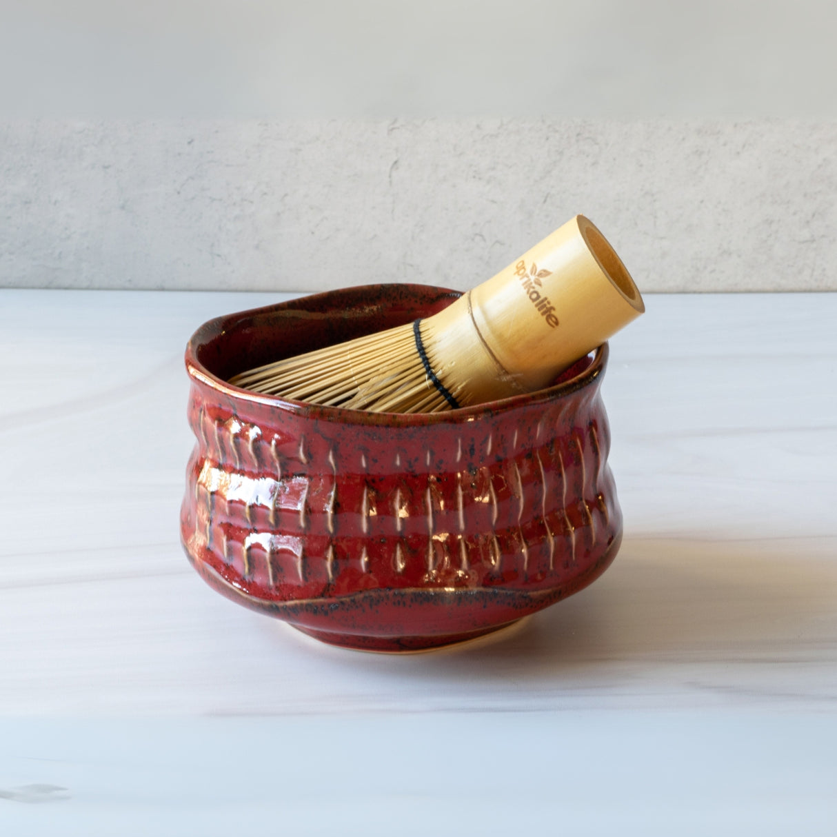 A Red Ceramic Matcha Bowl holds a Bamboo Matcha Whisk, placed elegantly on a light surface.