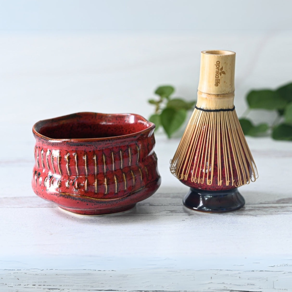 The Red Ceramic Meisen Matcha Bowl and bamboo whisk sit on a light wooden surface, framed elegantly by lush greenery.