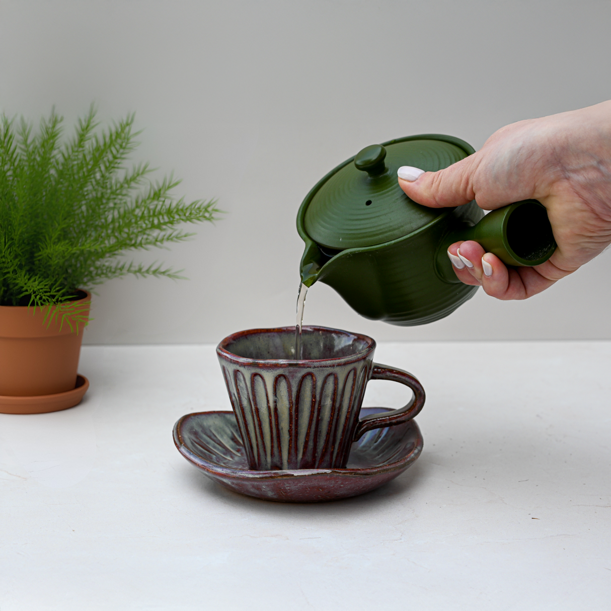 A hand pours tea from a green teapot into the Shibu Yunomi Japanese Tea Cup with Saucer beside a potted plant, creating a vintage atmosphere.