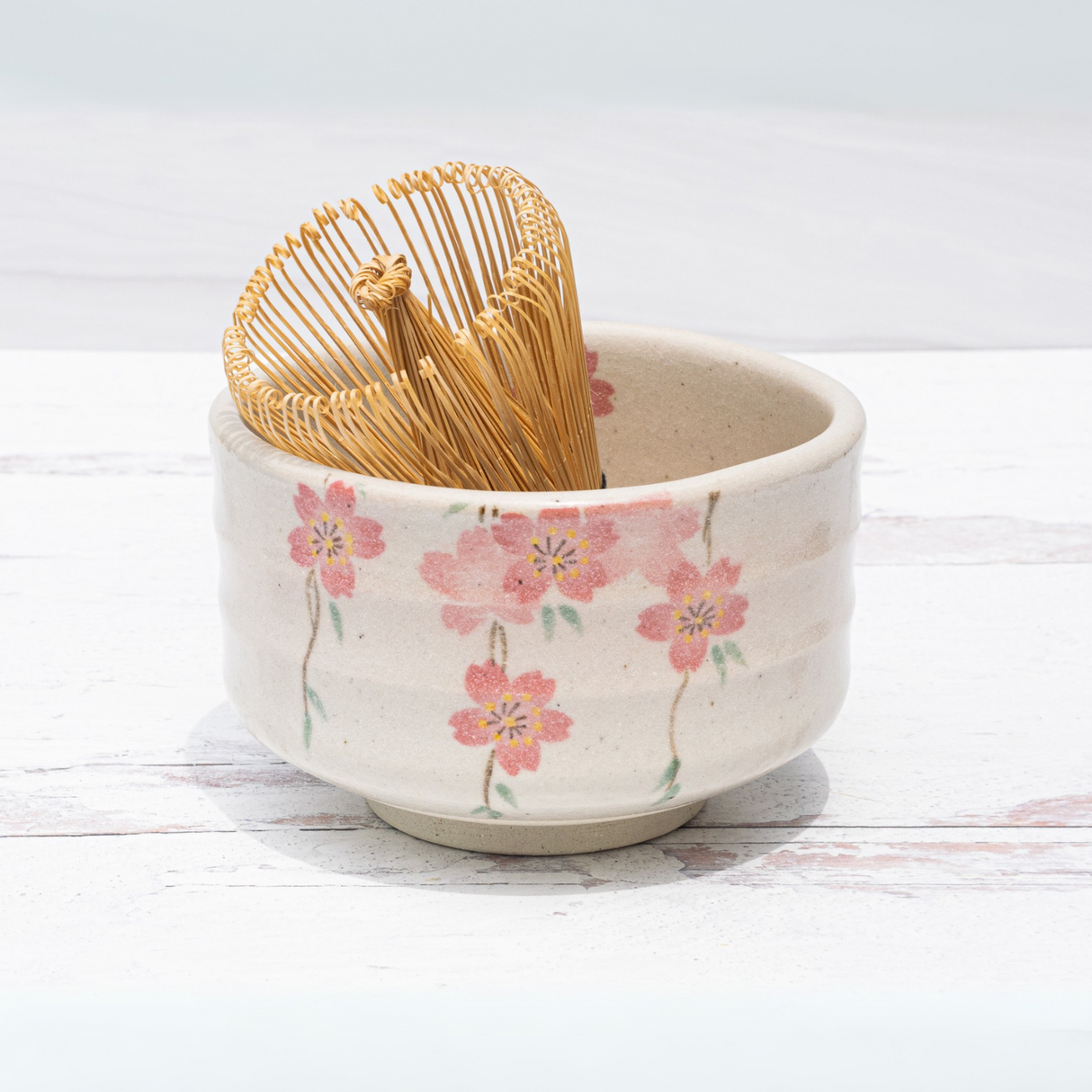 A bamboo matcha whisk rests in a ceramic bowl with pink cherry blossoms, part of the Pink Sakura Flowers Ceramic Matcha Bowl, Bamboo Matcha Whisk and Whisk Holder Set—beautiful teaware inspired by traditional Japanese pottery.