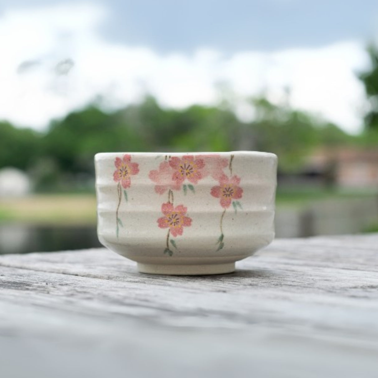 The Pink Sakura Flowers Ceramic Matcha Bowl with bamboo whisk and holder set, inspired by traditional Japanese pottery, sits on a wooden surface outdoors—perfect for enjoying matcha amidst lush greenery.