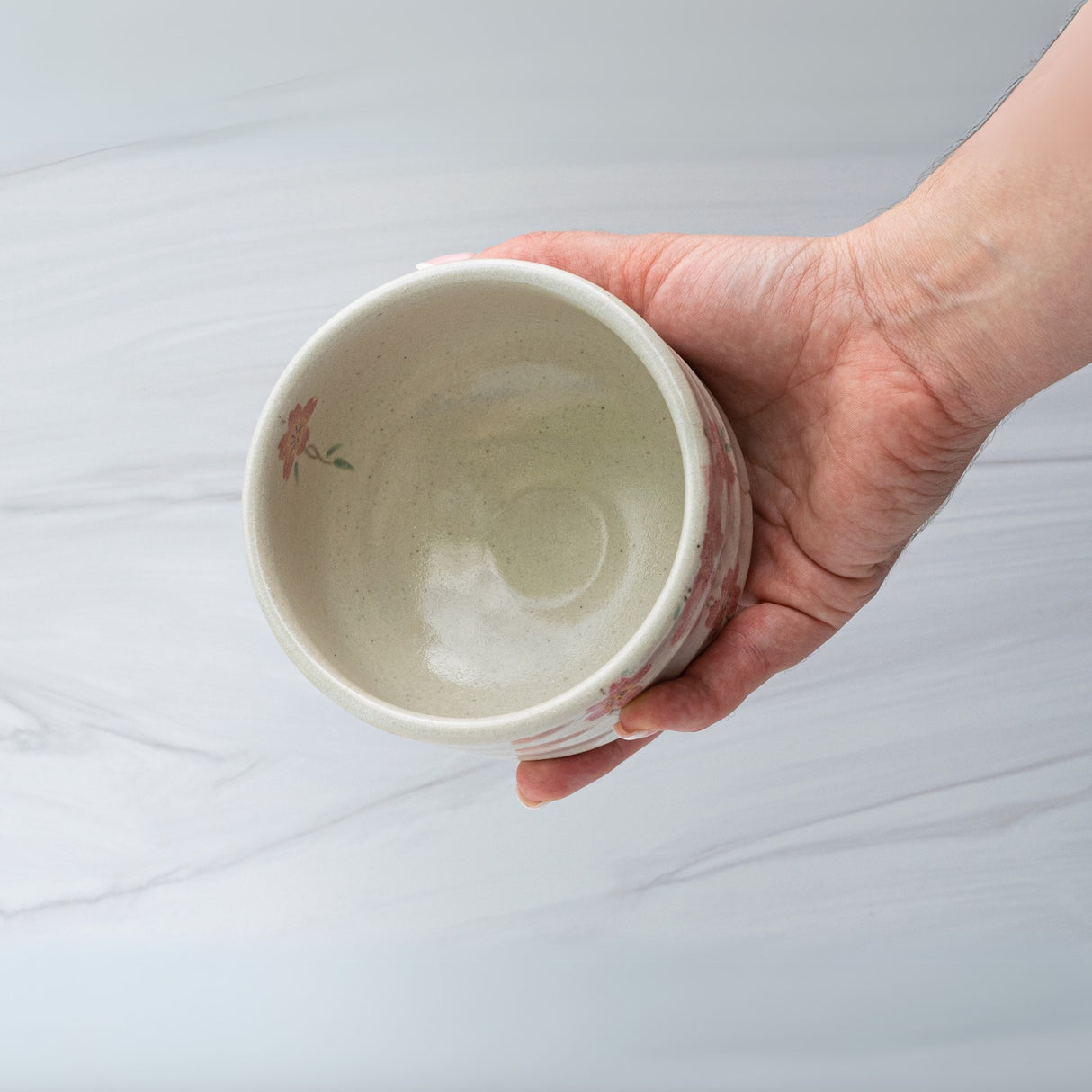 A hand holds the Pink Sakura Flowers Ceramic Matcha Bowl, part of a set with a bamboo matcha whisk and whisk holder, featuring delicate floral designs inspired by traditional Japanese teaware against a light background.