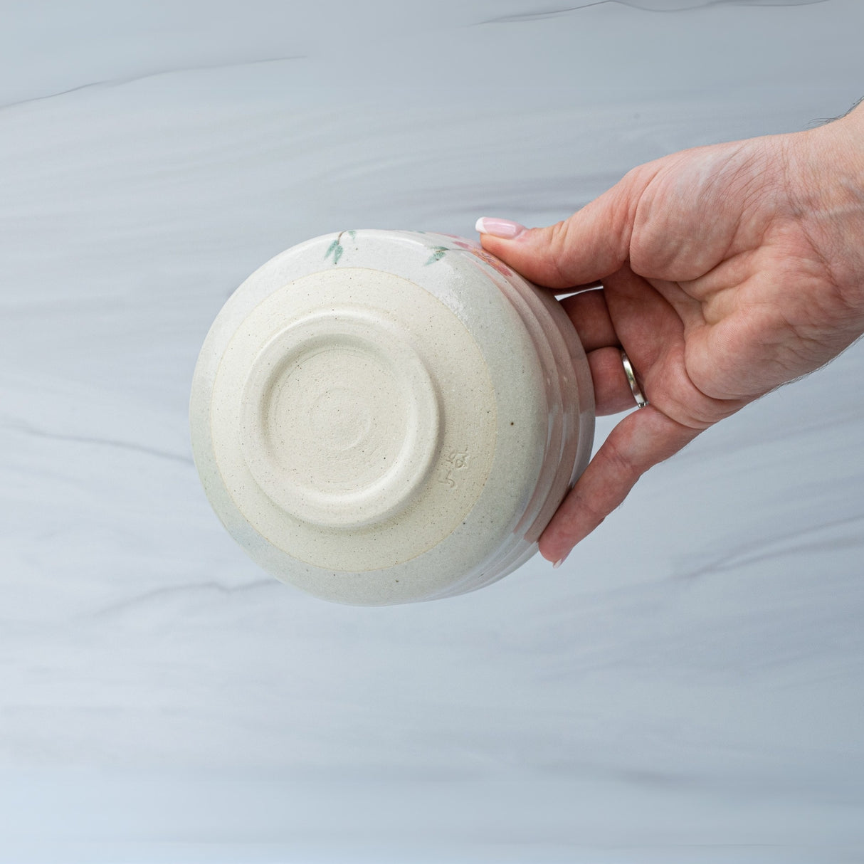 A hand holds an upside-down Pink Sakura Flowers Ceramic Matcha Bowl from the Bamboo Matcha Whisk and Whisk Holder Set against a light gray marbled background, highlighting the charm of traditional Japanese pottery.