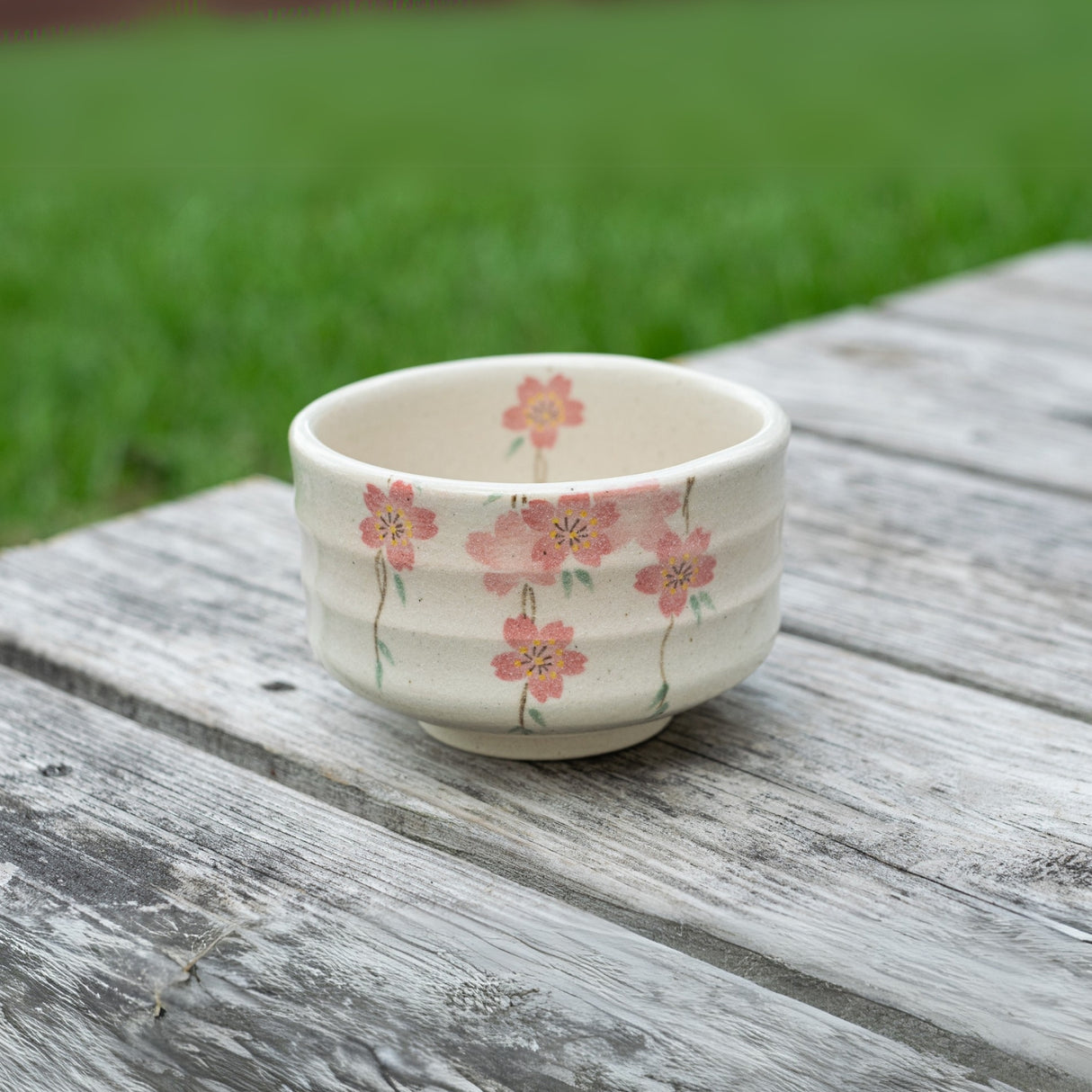 A Pink Sakura Flowers Ceramic Matcha Bowl from the Organic Japanese Matcha Set, featuring pink floral designs, rests on a weathered wooden surface outdoors, capturing the refined elegance of traditional Japanese pottery.