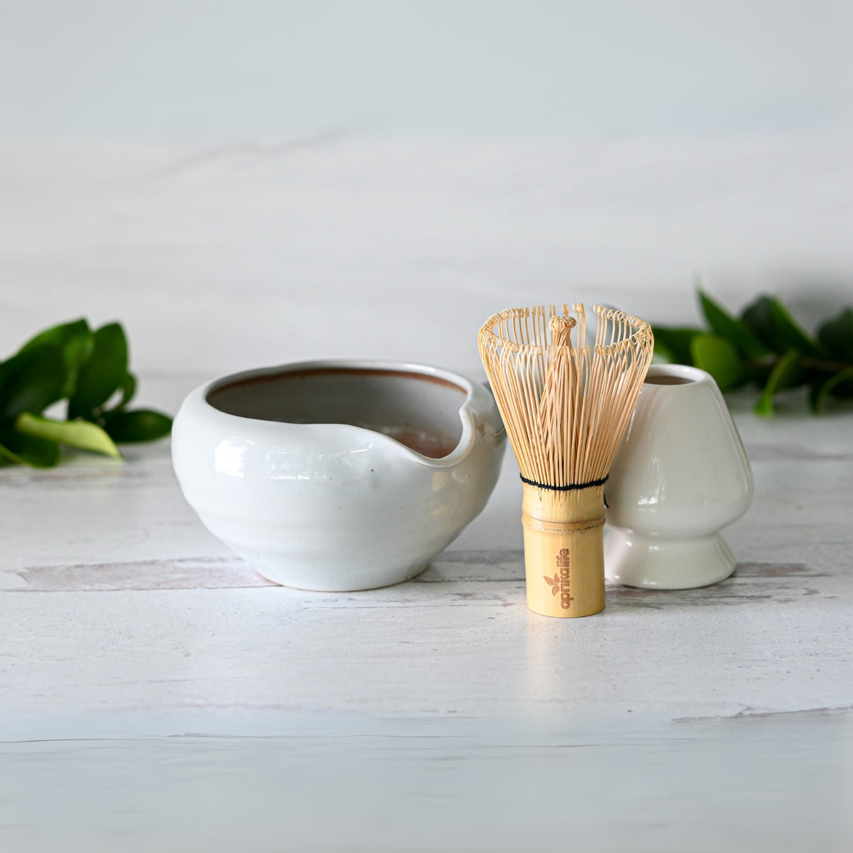 The Gray Spouted Ceramic Matcha Bowl with Textured Handle, Bamboo Matcha Whisk, and Whisk Holder from the Organic Japanese Matcha Set sits on a light wood surface, surrounded by green leaves.