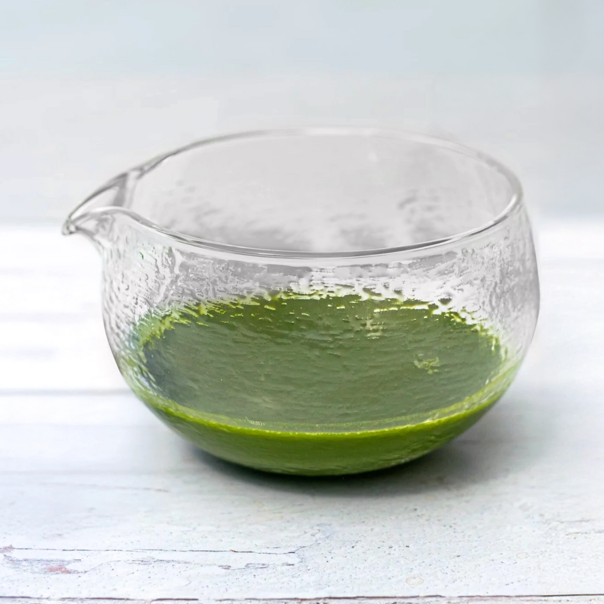 The Textured Spouted Glass Matcha Bowl Pattern 1, holding a small amount of green matcha, sits on a light wooden surface, showcasing minimalist teaware elegance.