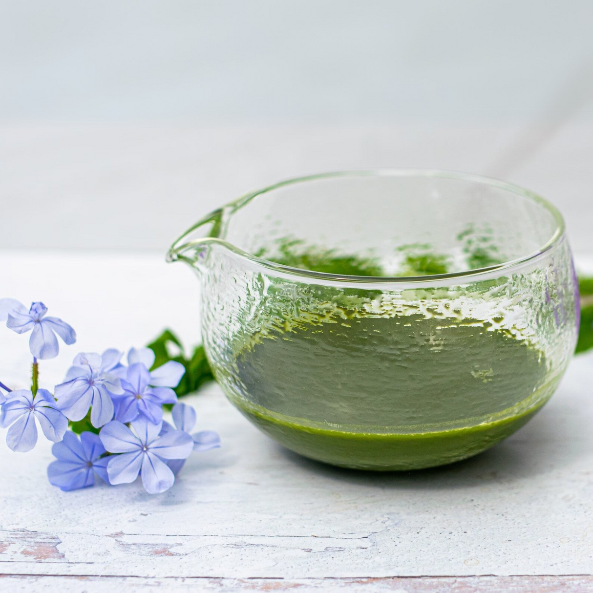 The Textured Spouted Glass Matcha Bowl Pattern 1, filled with green matcha, is placed next to light purple flowers on a white wooden surface.