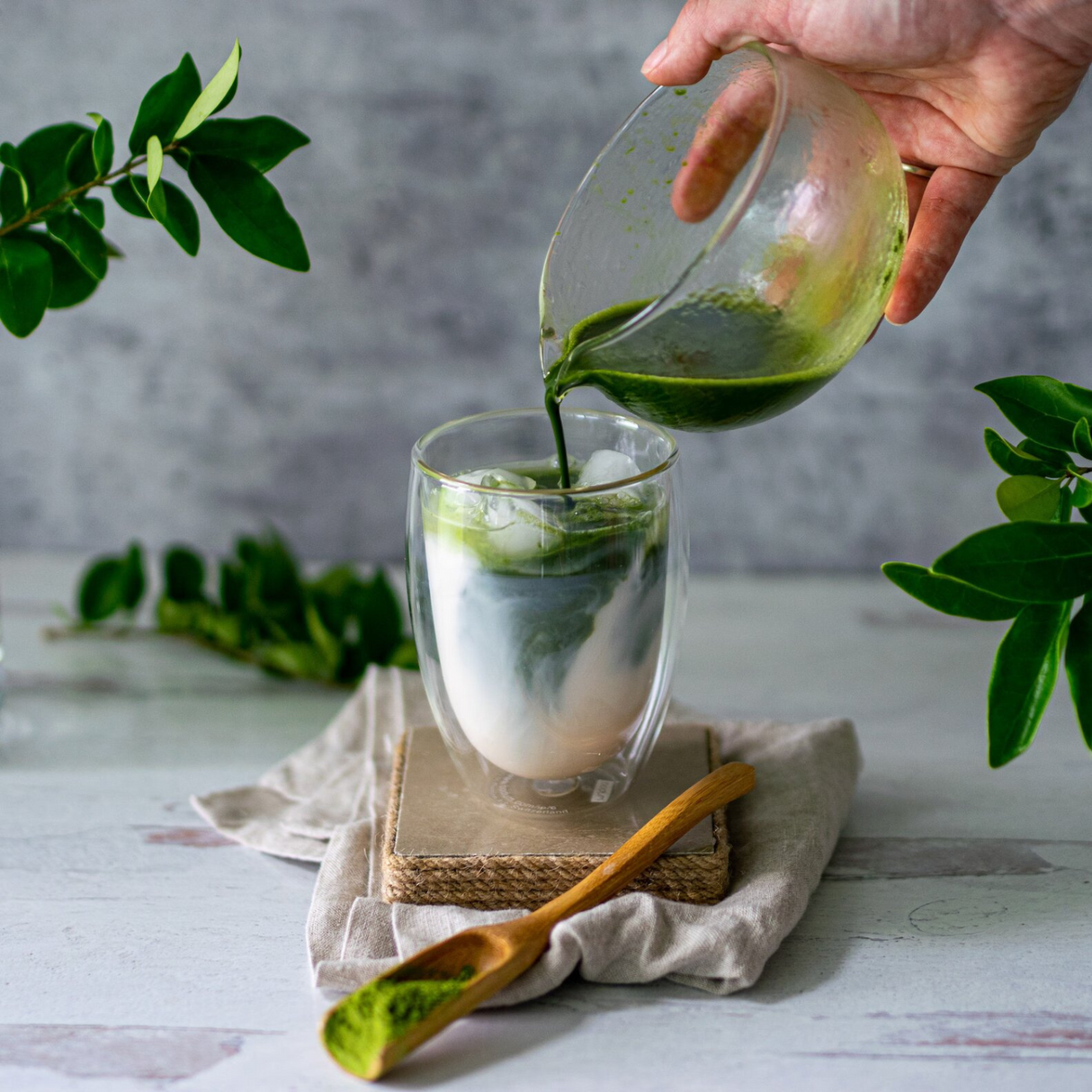 A hand pours green matcha from the Textured Spouted Glass Matcha Bowl Pattern 1 into a glass of milk on a table, with textured leaves and a wooden spoon nearby.