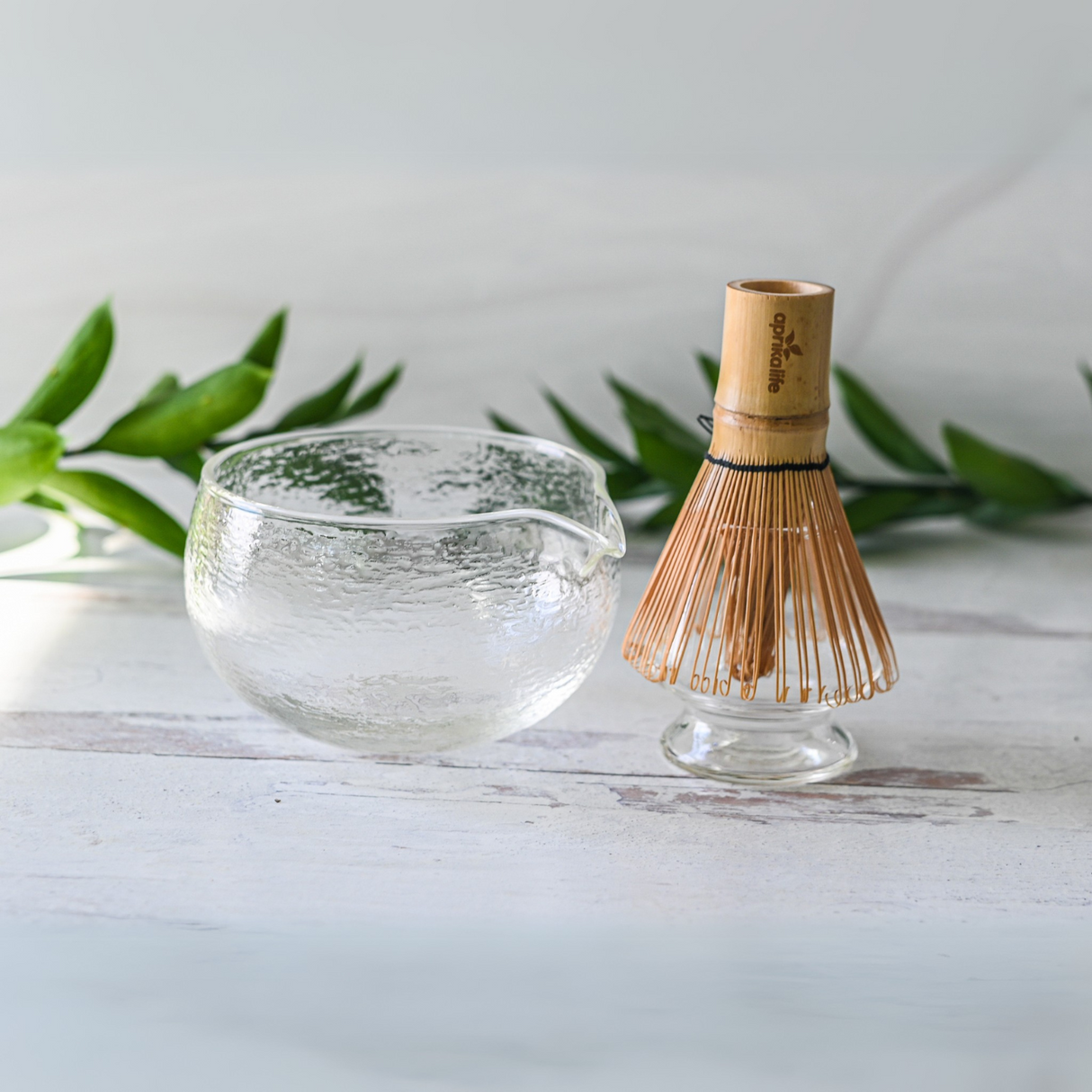 A Textured Spouted Glass Matcha Bowl Pattern 1 and a bamboo whisk rest on a white surface with green leaves in the background.