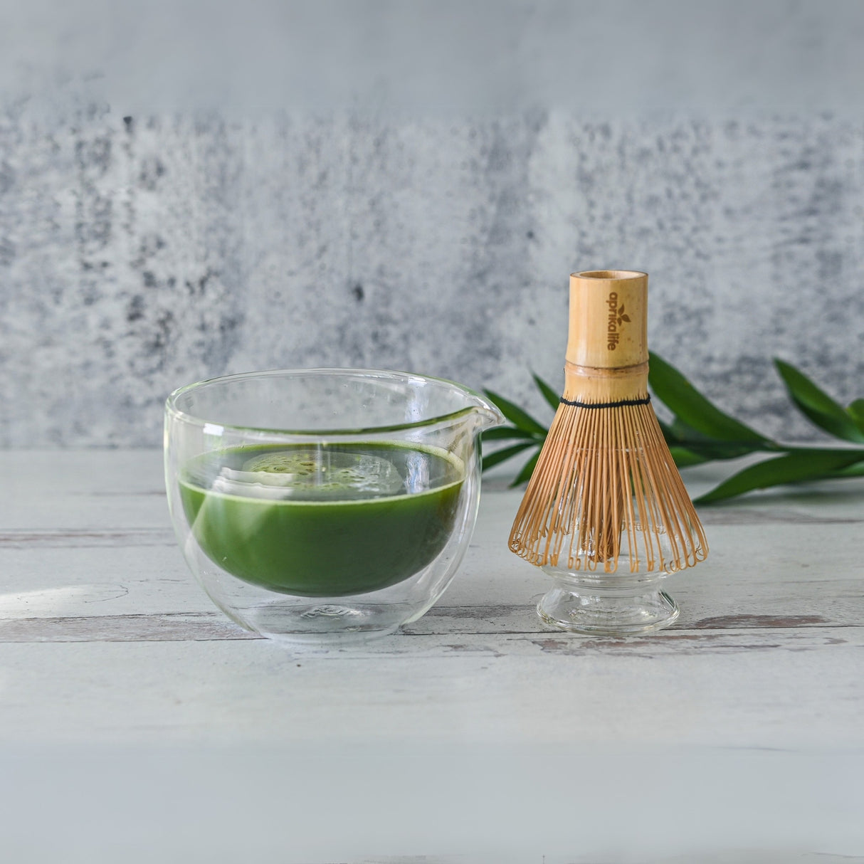 The Double Sided Glass Matcha Bowl with Spout holds vibrant green tea, complemented by a Bamboo Matcha Whisk and Whisk Holder, all resting on a light wooden surface.