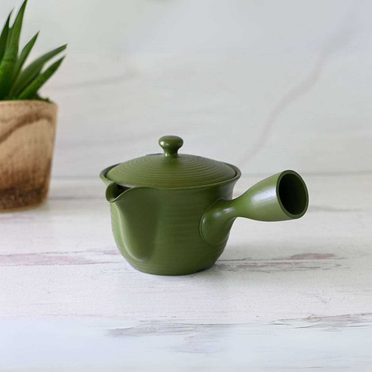 The Handcrafted Japanese Kyusu Teapot with Filters (380ml) in green, adorned with a side handle, gracefully rests on a white surface next to a potted plant. Its timeless design echoes Fujita Tokuta's style, adding elegance and tradition to any tea experience.