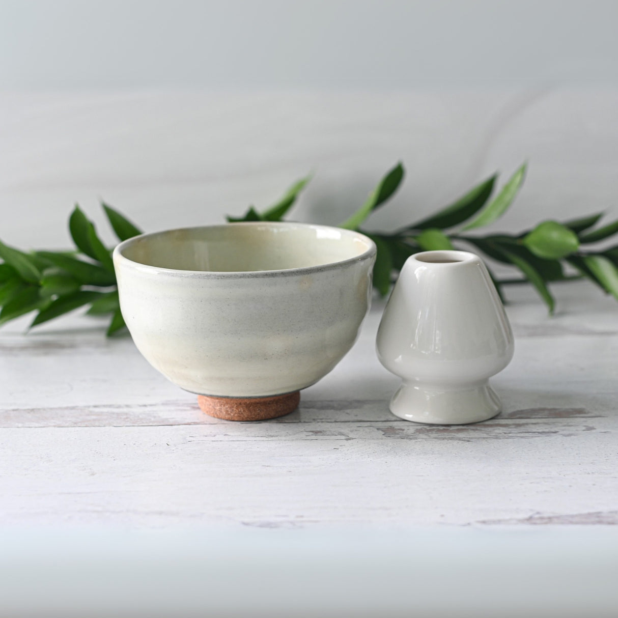 The White Kohiki Ceramic Matcha Bowl rests on a light wooden surface, accompanied by a small vase crafted with Minoyaki techniques, while green foliage sets a serene background.