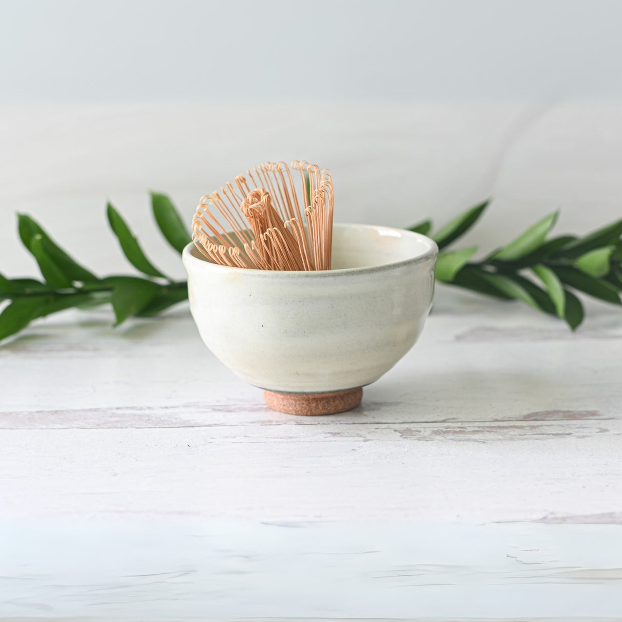 Experience the elegance of the White Kohiki Ceramic Matcha Bowl, crafted with traditional Minoyaki techniques. A bamboo matcha whisk elegantly rests inside, set against a light backdrop accented with vibrant green leaves.