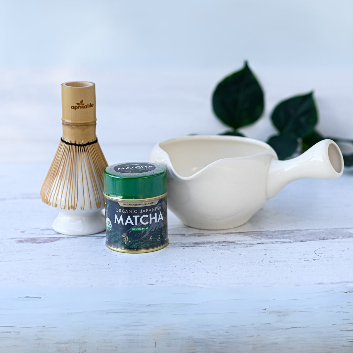 The White Ceramic Matcha Bowl with Spout, alongside a matcha tin and bamboo whisk on a white surface, surrounded by greenery, forms the perfect ensemble for a Japanese tea ceremony or as a thoughtful housewarming gift.