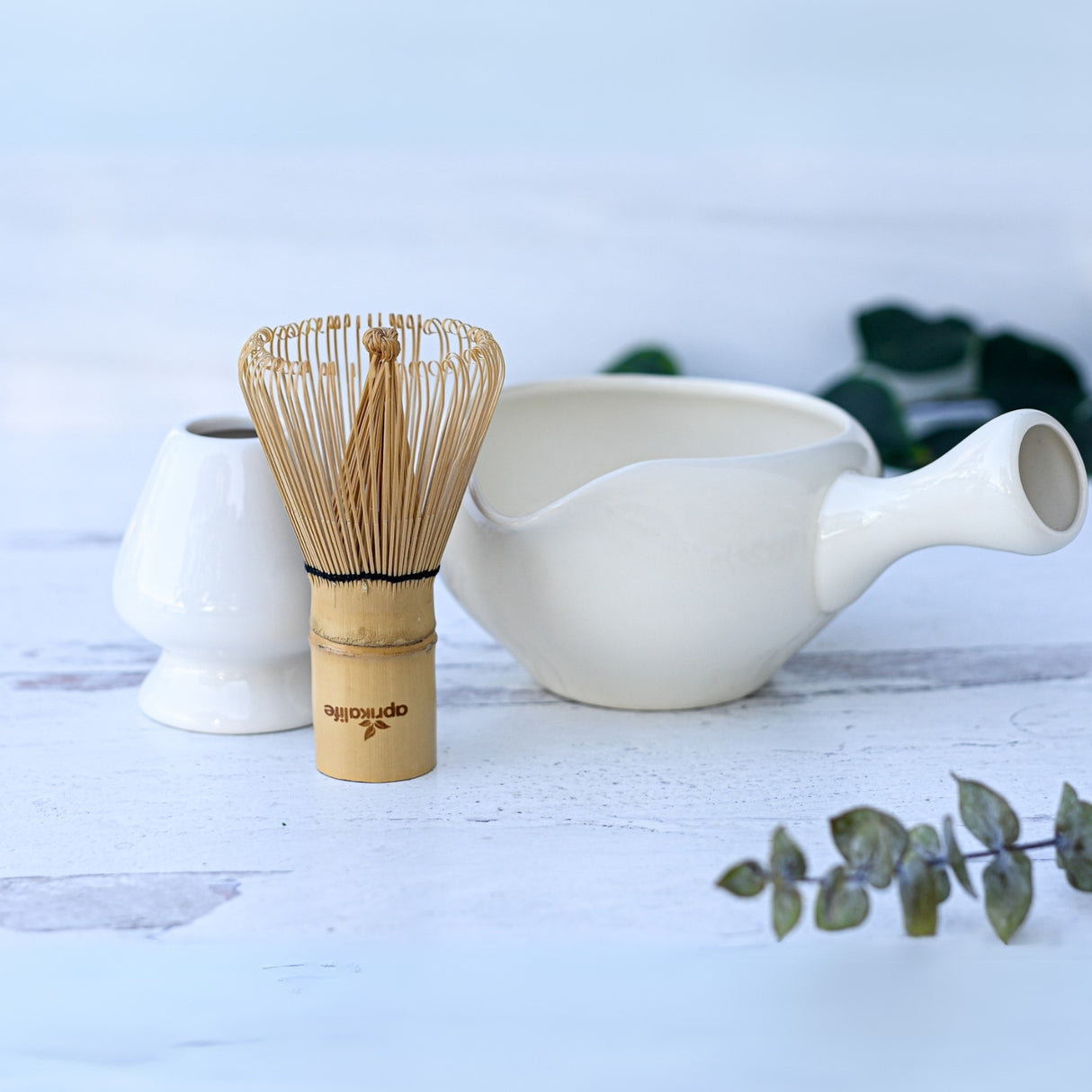 A bamboo matcha whisk sits elegantly beside a White Ceramic Matcha Bowl with Spout, framed by lush greenery. Perfect for a Japanese tea ceremony or as a thoughtful housewarming gift.