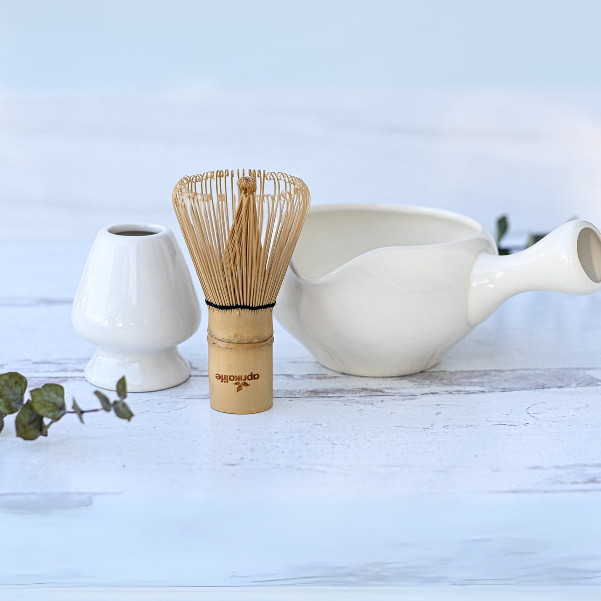A tea whisk and holder accompany a white teapot on a light wooden surface, paired with our White Ceramic Matcha Bowl with Spout and a nearby leaf—an ideal housewarming gift for those who appreciate the elegance of the Japanese tea ceremony.