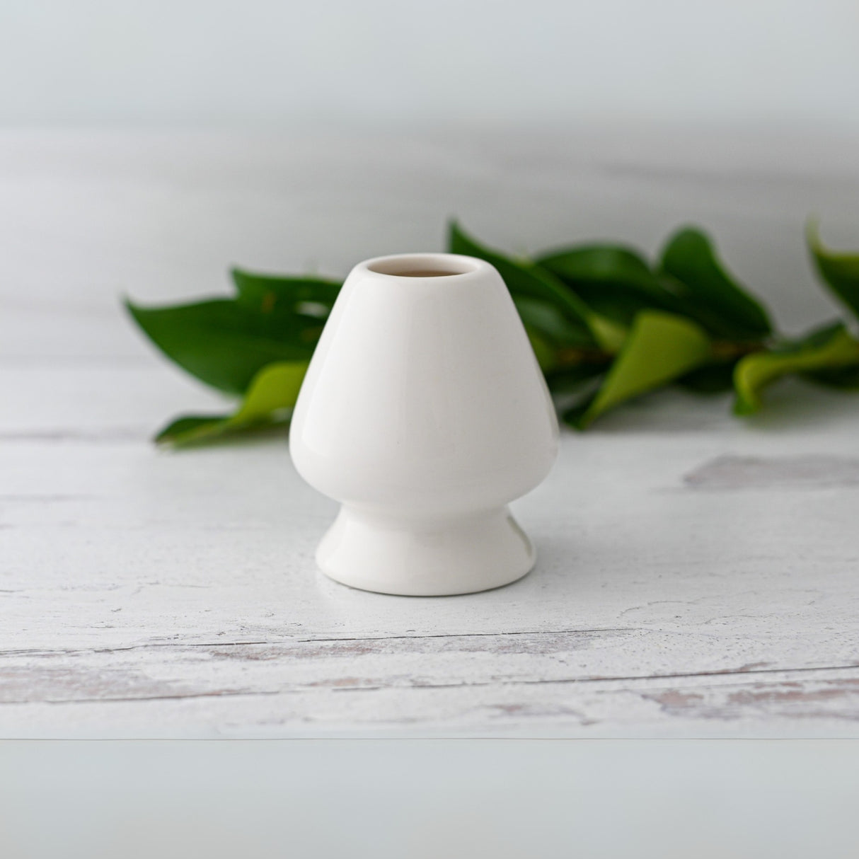 A White Matcha Whisk Holder from the Bamboo Whisk Set stands elegantly against a light background, with softly blurred green leaves in the distance.